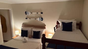 Quarto de hotel com duas camas, cada uma com cabeceira de madeira, uma mesinha de cabeceira com abajur e um porta-toalhas fixado na parede.
