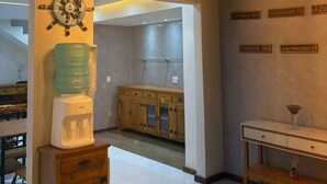 A modern interior with a water dispenser, wooden cabinets, a table with a glass, and hanging light fixtures. - Suítes Brisa do Mar (Búzios)