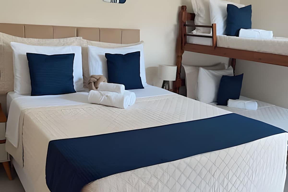 A hotel room with a bunk bed, a large bed with white bedding and blue pillows, and a bedside table with a lamp.