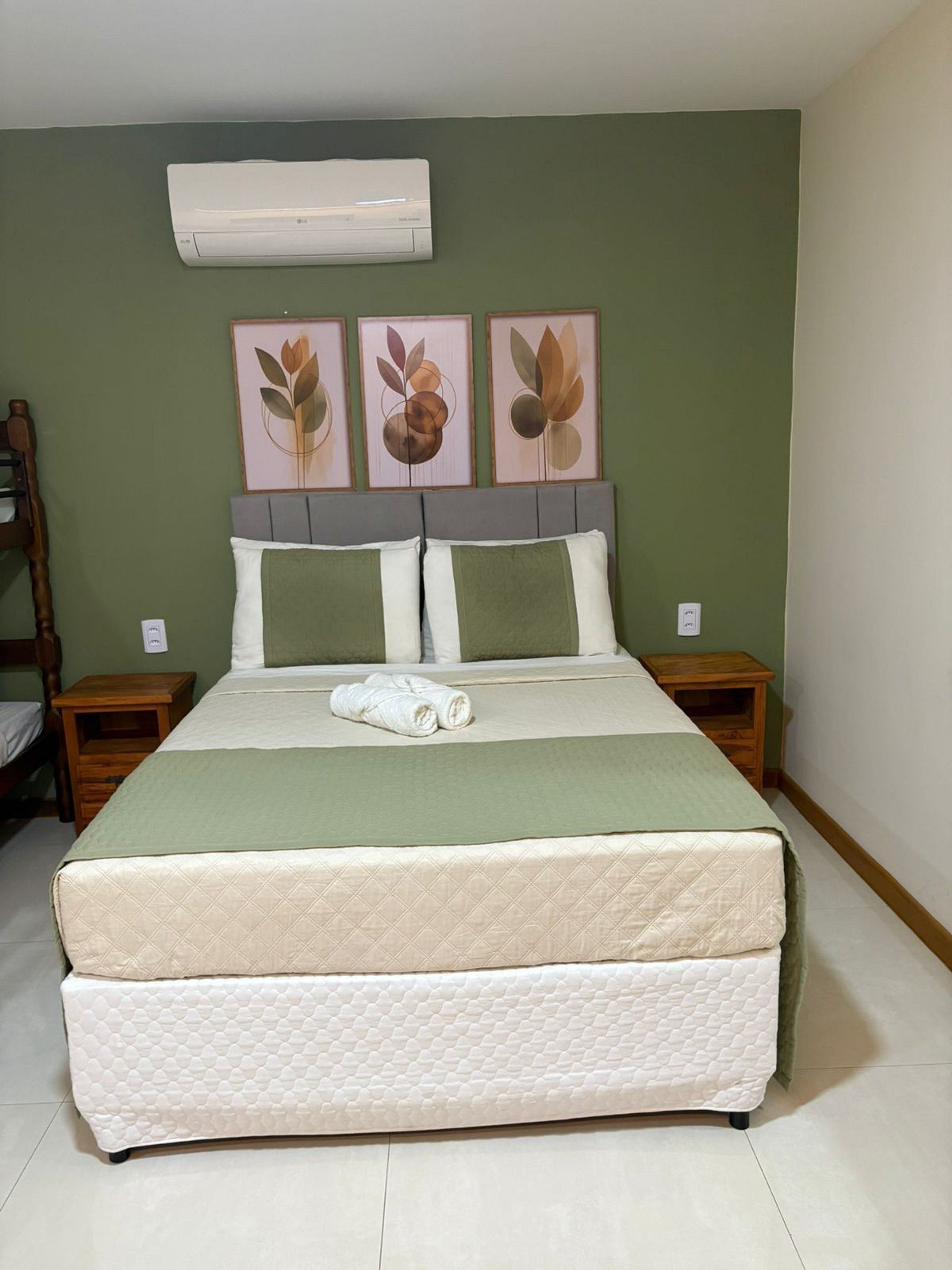 Una cama bien hecha con edredón verde y blanco, dos almohadas y una toalla enrollada sobre la cama. Hay dos mesitas de noche con lámparas, aire acondicionado de pared y tres cuadros florales enmarcados sobre la cama.