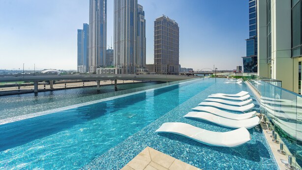 A rooftop infinity pool with clear blue water, surrounded by modern buildings and a bridge in the background.