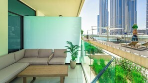 A modern living room with a sofa, a coffee table, and a potted plant. A balcony overlooks a pool area with lounge chairs and a person walking.