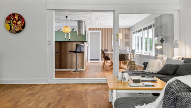 A modern living room with a wooden floor, a sofa, a coffee table with books, and a dining area with a table and chairs. - 3 bedroom beautiful home in Gilleleje (Gilleleje)