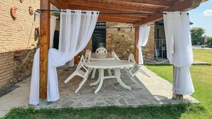 A covered patio with a stone table and chairs, white curtains, and a brick wall.