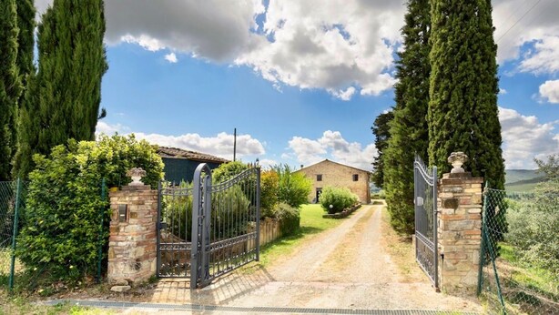 A rural property with a gravel driveway leading to a stone gate, surrounded by tall cypress trees and a stone building with a tiled roof.