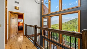 Una escalera de madera conduce a un balcón con ventanas amplias que ofrecen vistas de una ladera boscosa.