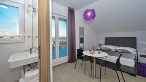 Un hotel moderno con balcón, una mesa redonda de comedor, una cama con ropa de cama gris y un ventanal con vistas al mar.