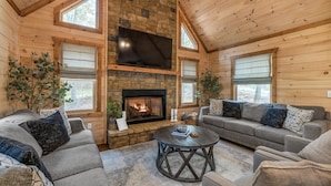 A cozy log cabin interior with a stone fireplace, two sofas, a round coffee table, and large windows. - Large Family Getaway Cabin w/Hot Tub + Game Room - Minutes to Blue Ridge (Blue Ridge)