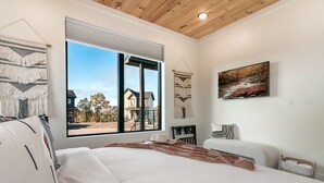 Une chambre moderne avec une grande fenêtre, un lit recouvert de linge de lit blanc, un plaid marron et un tableau représentant une scène de forêt.