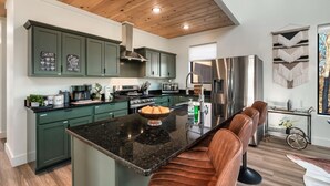 Une cuisine moderne avec des meubles de couleur verte, un îlot central en granit noir, des appareils électroménagers en acier inoxydable et un coin salon avec des fauteuils en cuir.