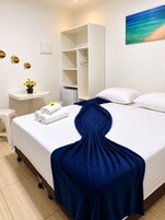 Quarto de hotel com cama, guarda-roupa branco, secretária pequena com cadeira e prateleira na parede.