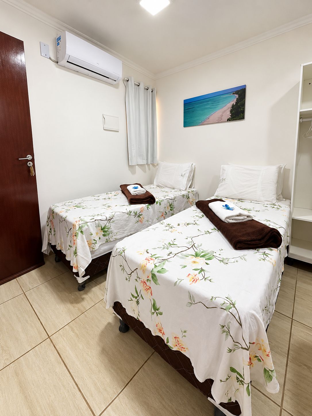 Habitación de hotel con dos camas, una puerta, un equipo de aire acondicionado, un cuadro de playa y una cortina blanca.