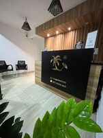 Balcão de recepção com painel de madeira ao fundo, bancada preta e logotipo de palmeira. Duas cadeiras pretas estão em frente ao balcão, e uma planta é visível em primeiro plano.