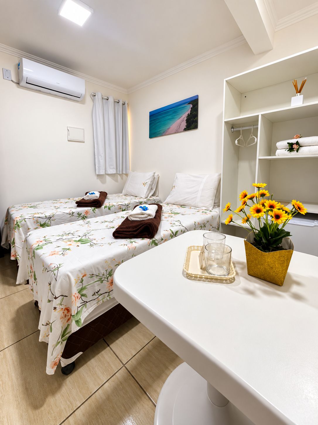 A hotel room with two beds, a small table with a glass and a vase of flowers, a painting of a beach, and a shelf with towels and a plant.
