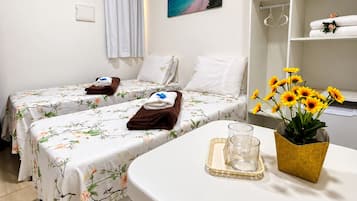 A hotel room with two beds, a small table with a glass and a vase of flowers, a painting of a beach, and a shelf with towels and a plant.