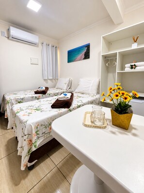 Habitación de hotel con dos camas, una mesita con un vaso y un jarrón de flores, un cuadro de una playa y un estante con toallas y una planta.