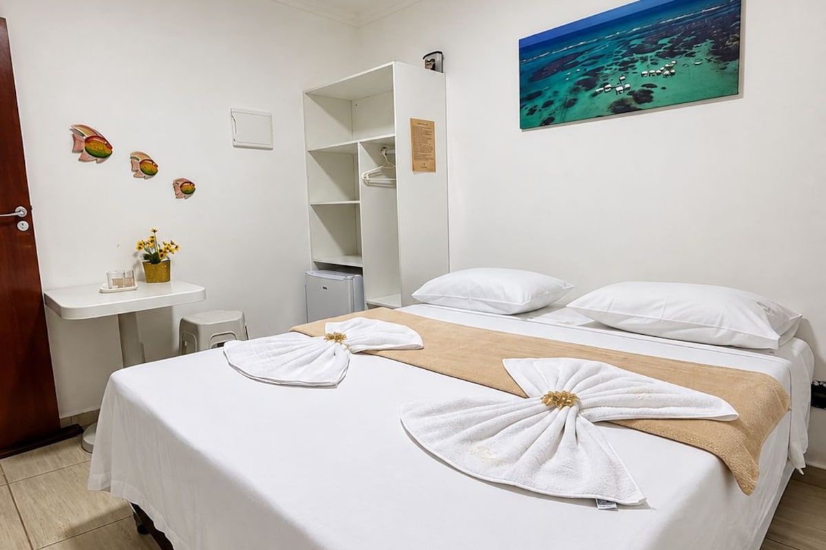 Una habitación de hotel con una cama, un escritorio, una silla, una mesita con un florero y un cuadro de una playa.