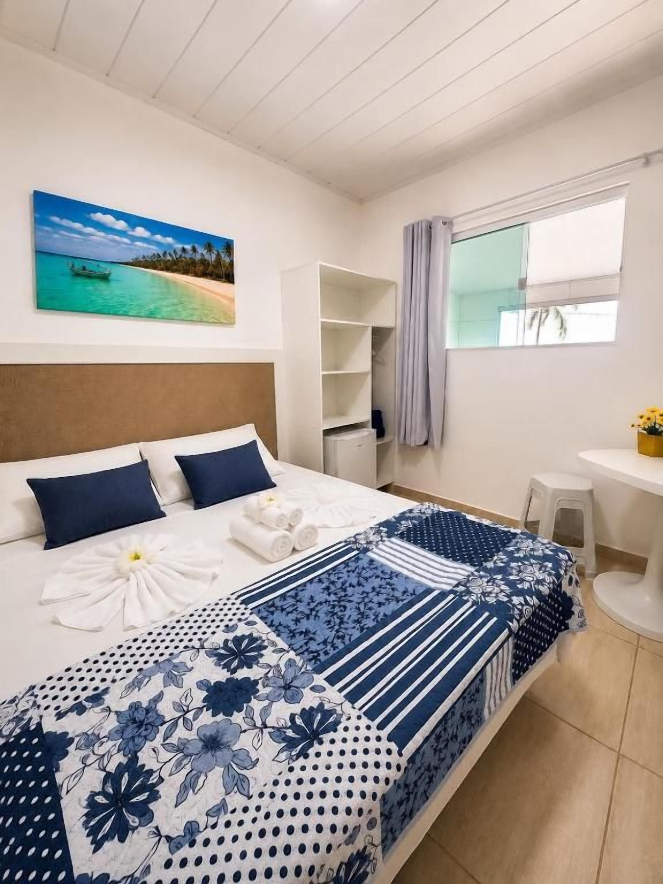 A hotel room with a bed, a painting of a beach, a window with curtains, a small table, and a white stool.