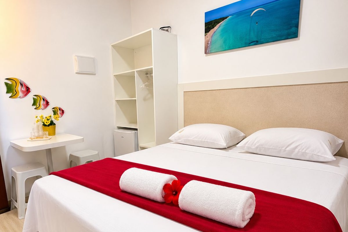 Una habitación de hotel con una cama, un detalle en rojo, un cuadro y una pequeña mesa con un florero.