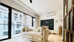 Een moderne woonkamer met een beige bank, grote ramen, een schoolbord met tekst en uitzicht op een gebouw.