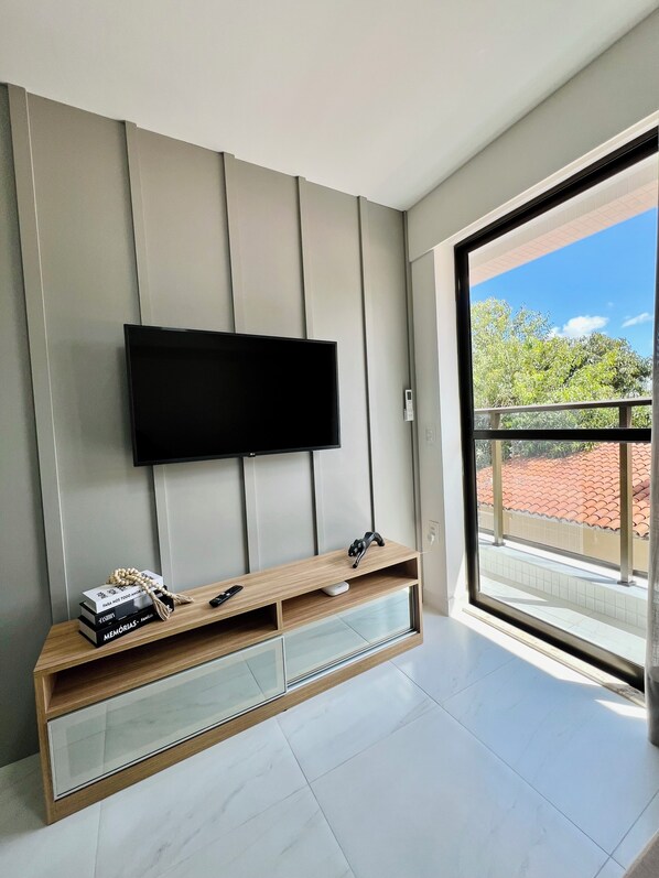 Ein modernes Wohnzimmer mit einem flachbildfernseher an der Wand, einer Holzunterhaltungseinheit darunter und einem großen Fenster mit Blick auf ein rotes Ziegeldach und Grünflächen.
