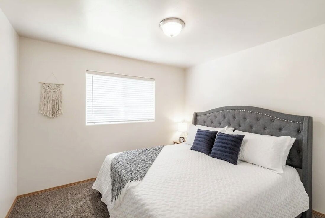 Um quarto com cama, mesa de cabeceira, um detalhe de macramé na parede e janela com persianas.