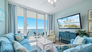 A living room with a large TV, a sofa, a coffee table, and a view of the ocean through large sliding doors.