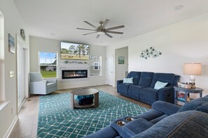 A living room with a fireplace, a ceiling fan, a blue couch, and a view of the outdoors. - Robert's Retreat | 5BR Marsh Views + Decks, Pools, Golf & Captain's Cove Amenities (Greenbackville)