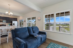 A modern living room with a blue sofa, dining area, kitchen, and large windows offering a view of the outdoors.