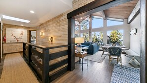 A rustic interior with wooden walls, a wooden railing, a dining area with a table and chairs, and a view of the outdoors through large windows.