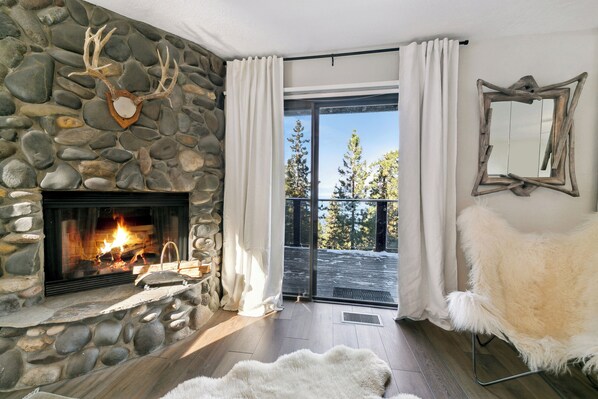 Una chimenea de piedra con un trofeo de venado, un sillón blanco y mullido, y vistas a árboles a través de un ventanal.