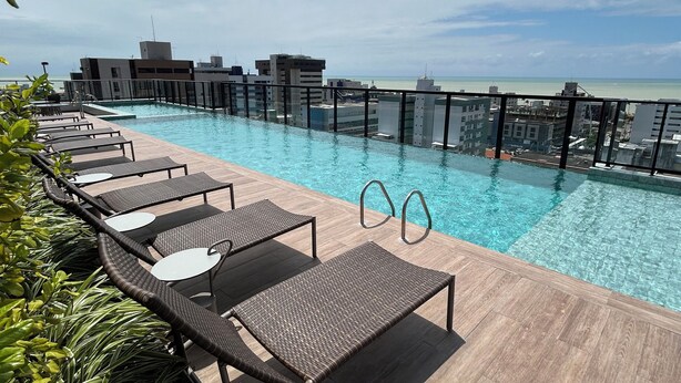 A rooftop pool with lounge chairs, a clear view of the ocean, and modern buildings in the background. - Melhores Flats (João Pessoa)