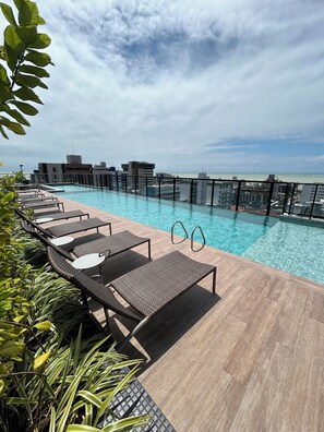 A rooftop pool with lounge chairs, a clear view of the ocean, and modern buildings in the background. - Melhores Flats (João Pessoa)
