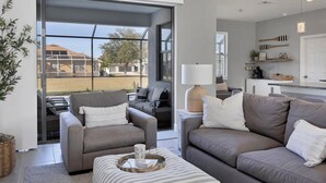 A modern living room with a grey sofa, a striped ottoman, and a coffee table. There is a sliding glass door leading to an outdoor area with a view of houses. - Punta Gorda Private Pool Vacation Home (Punta Gorda)