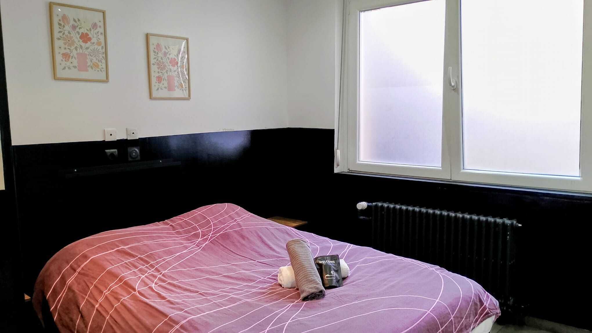 Een bed met een roze sprei, een opgerolde handdoek en een welkomstpakket erop. Daarboven hangen twee florale kunstwerken in lijsten aan de muur.