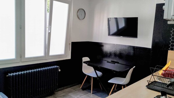 Una pequeña cocina americana con cubierta negra, dos sillas blancas, una mesa negra, un reloj en la pared y una ventana con persianas.