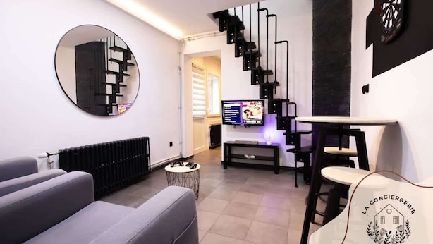 Un salon moderne avec un canapé gris, un miroir rond, un escalier noir, un téléviseur à écran plat et une table ornée d’un logo.