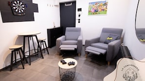 Un salon moderne avec un jeu de fléchettes, un fauteuil gris, une petite table ronde avec deux manettes de jeu et un logo sur le mur.