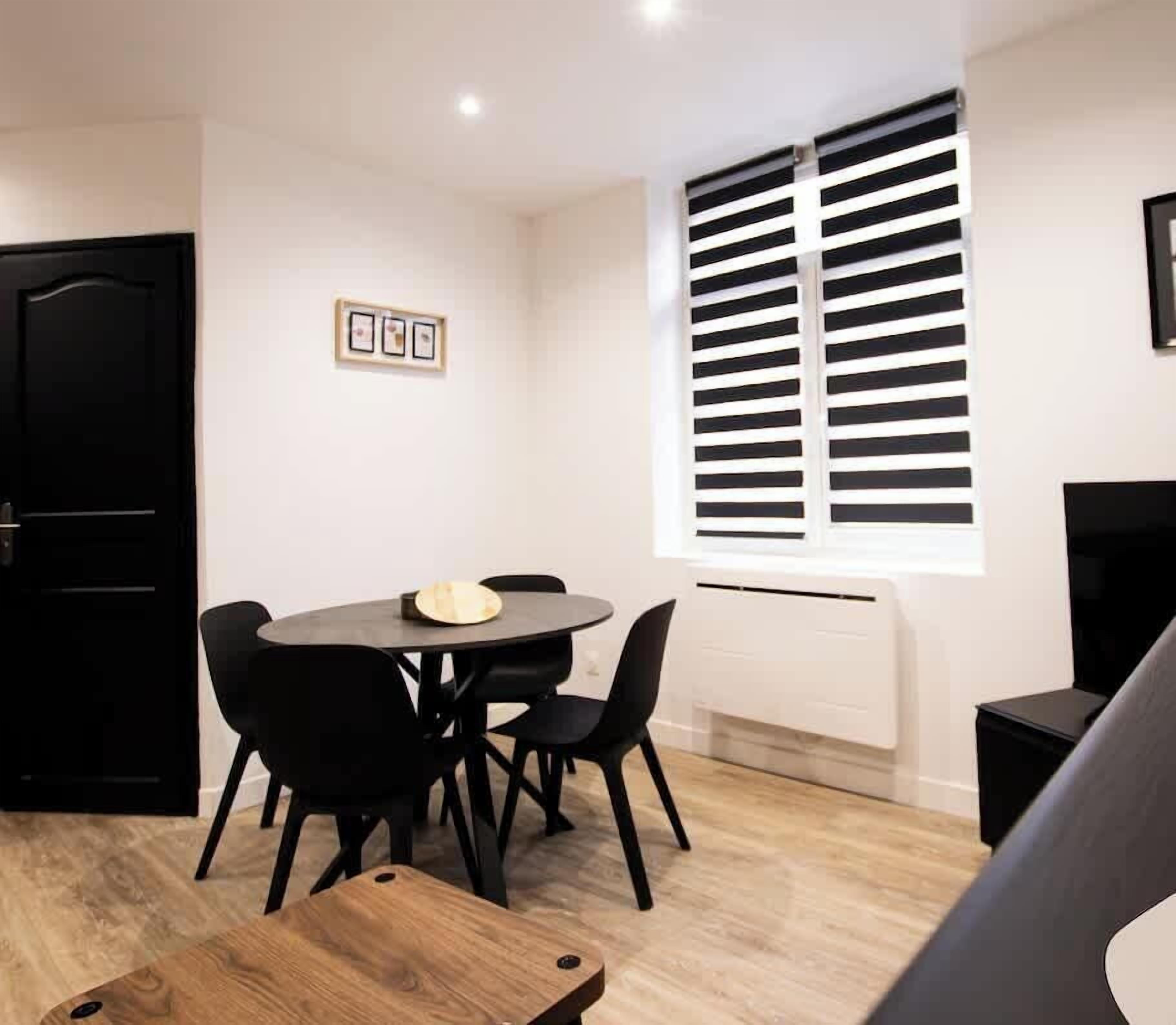 Una habitación moderna y minimalista con una mesa redonda y cuatro sillas, una puerta negra, un ventanal con persianas horizontales y un radiador blanco.