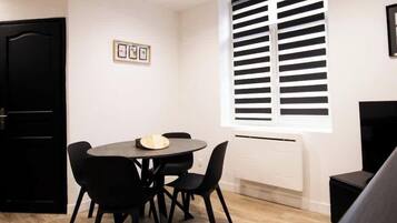 Una habitación moderna y minimalista con una mesa redonda y cuatro sillas, una puerta negra, un ventanal con persianas horizontales y un radiador blanco.