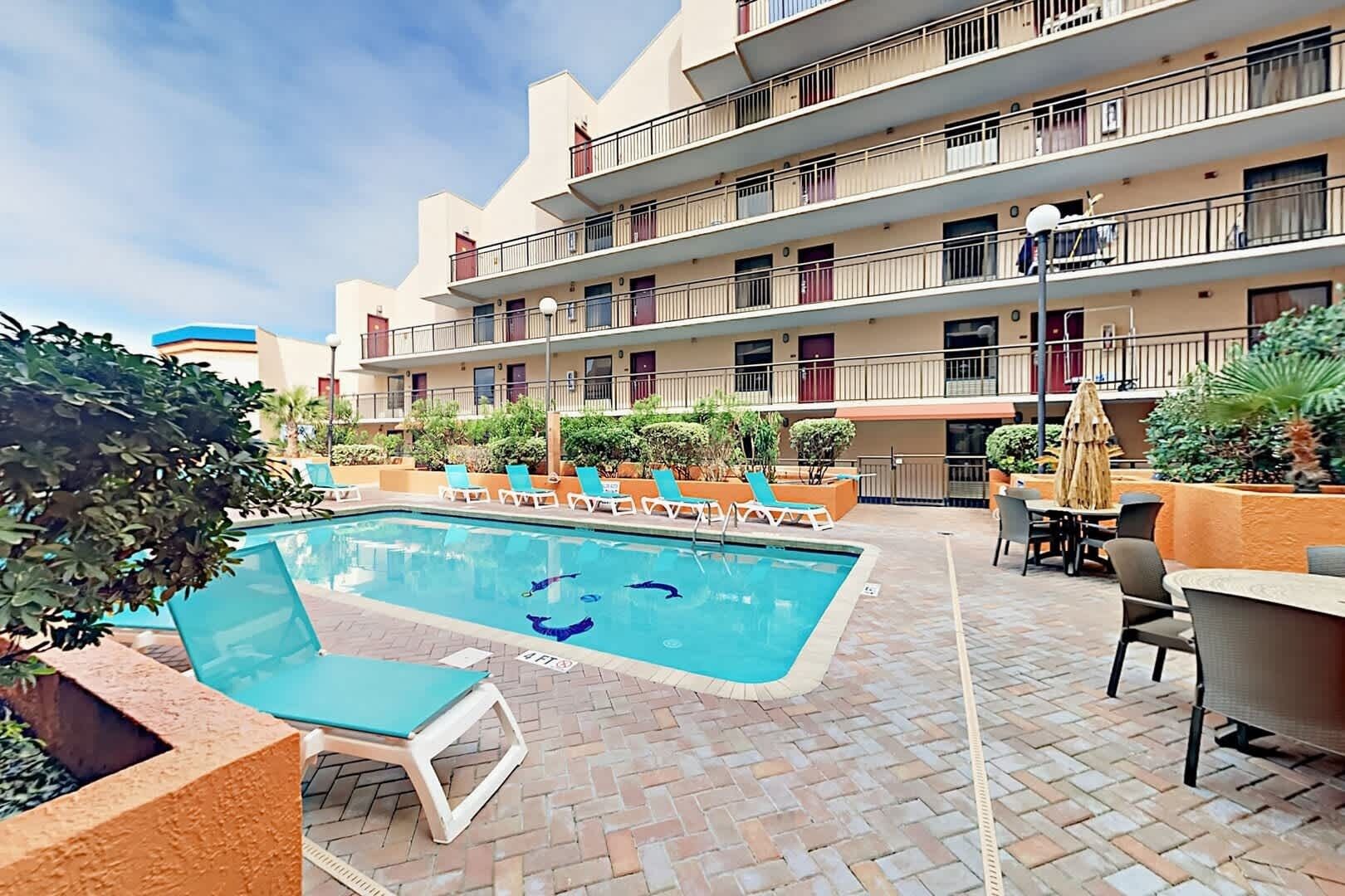 A hotel pool area with lounge chairs, a swimming pool, and a multi-story building with balconies.