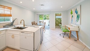 A modern kitchen with a dishwasher, a sink, and a dining area with chairs. There is a window with blinds and a door leading outside.