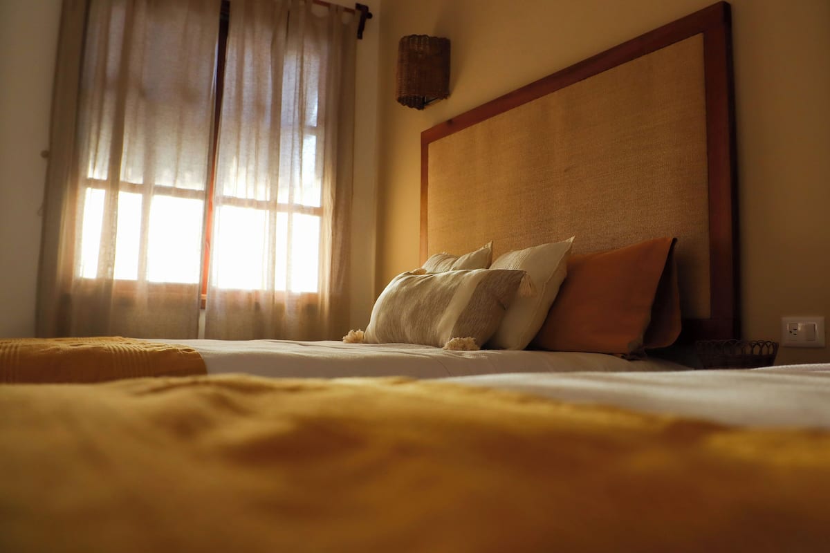 Un lit avec une tête de lit en bois, une fenêtre avec des rideaux légers et un luminaire fixé au mur.