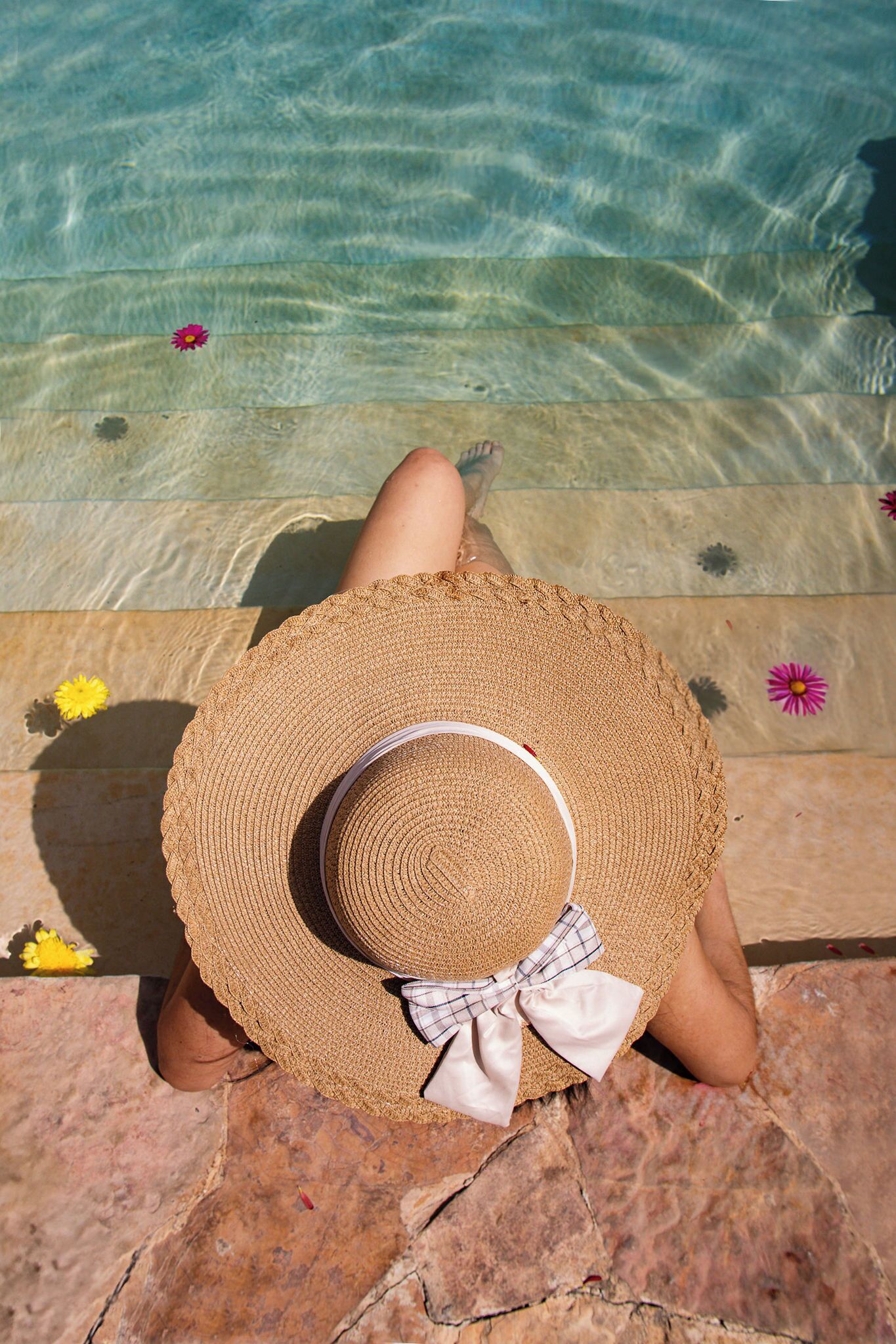 Una persona sentada junto a una piscina, con sombrero de paja y lazo, rodeada de flores.