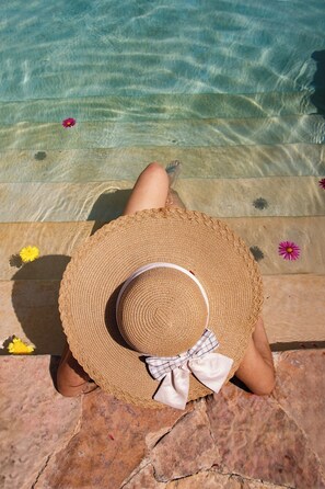 A person sitting by a pool with a straw hat and a bow, surrounded by flowers.
