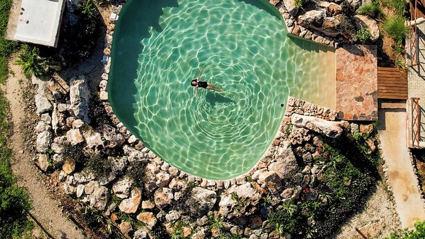 Ein Luftbild eines kreisförmigen Schwimmbeckens, umgeben von Felsen und Grünpflanzen, mit einer Person, die im Zentrum schwimmt.