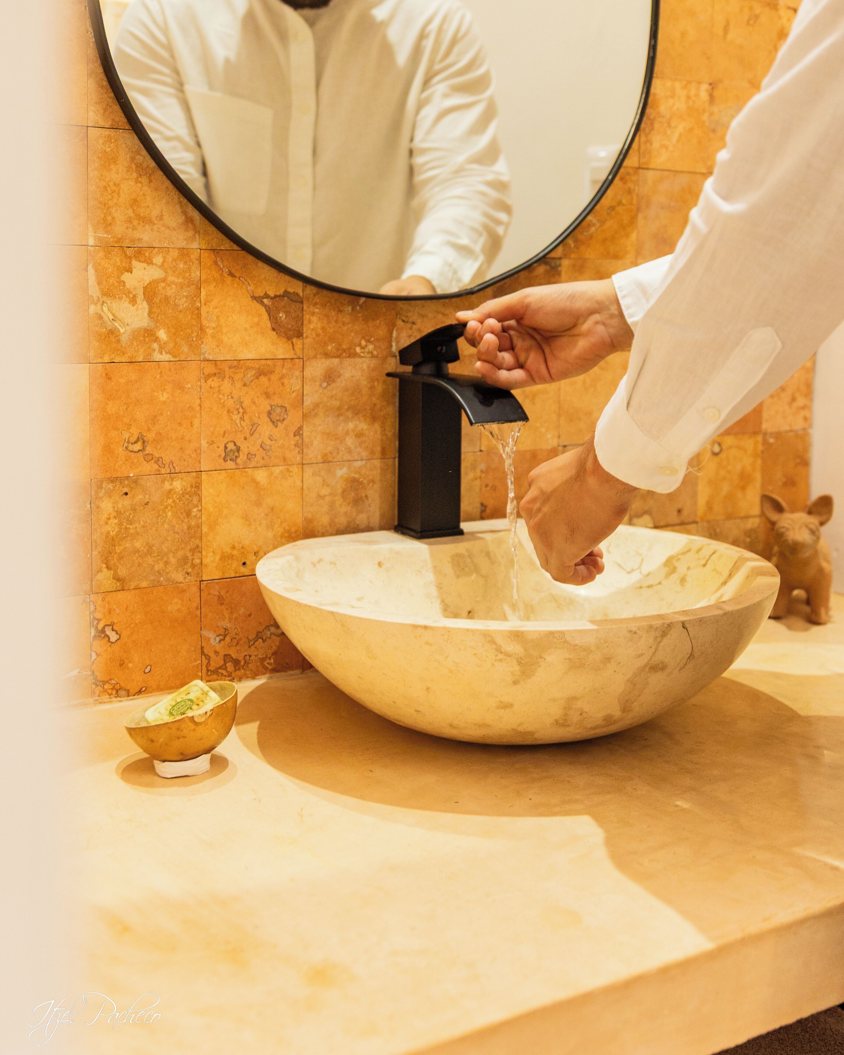 Un baño con un lavabo de mármol, un espejo redondo y alguien lavándose las manos en el grifo.