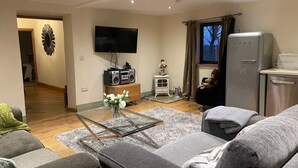 A living room with a sofa, a glass coffee table, a television, a radio, a decorative mirror, a small heater, and a refrigerator. - Meagill Cottage, Sleeps 4 With Hot Tub & Parking (Otley)