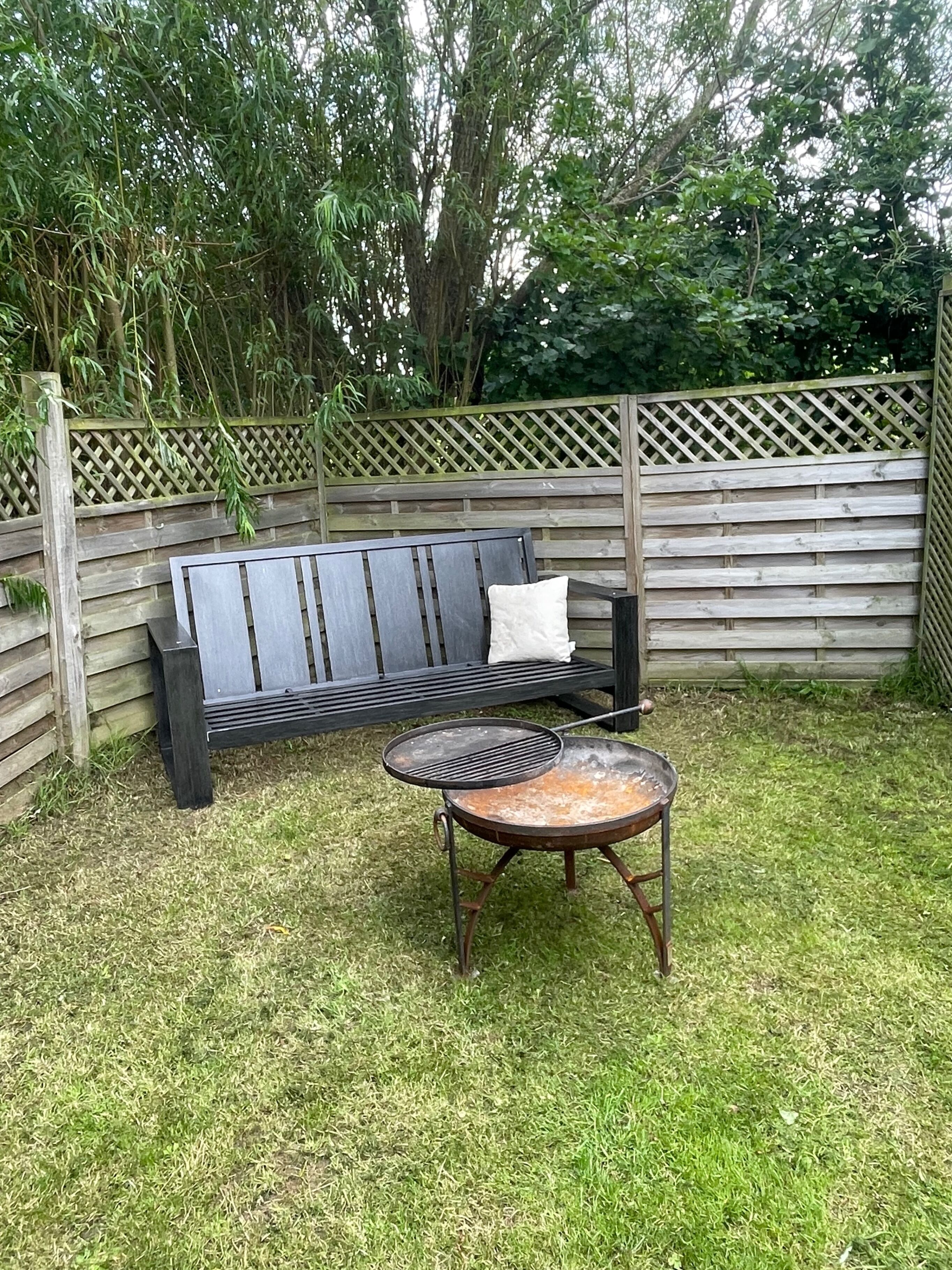A wooden fence, a black metal bench with a cushion, and a metal grill table in a grassy backyard.
