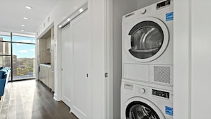 A laundry room with a front-loading washer and dryer, a large window with a city view, and a blue chair in the corner.